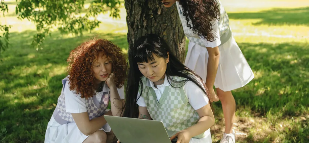 Tiga orang berkumpul di sekitar laptop di bawah pohon di taman yang terang.